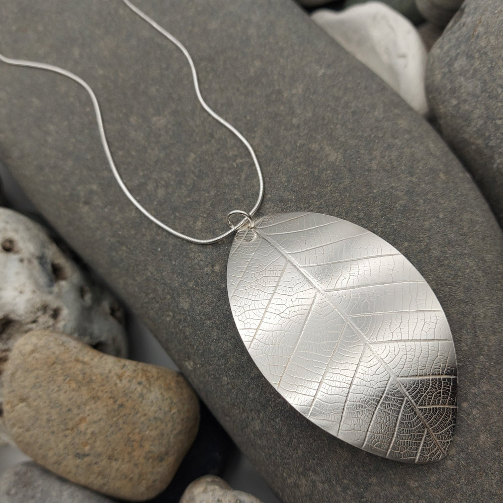Close up of Silver leaf-shaped and textured pendant on a chain lying on gray stones