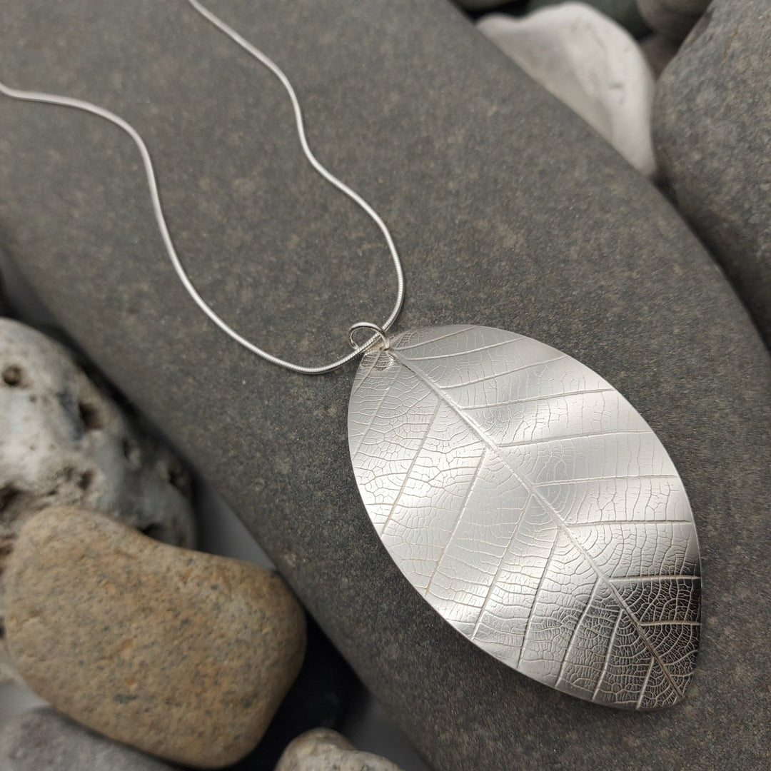 Close up of Silver leaf-shaped and textured pendant on a chain lying on gray stones