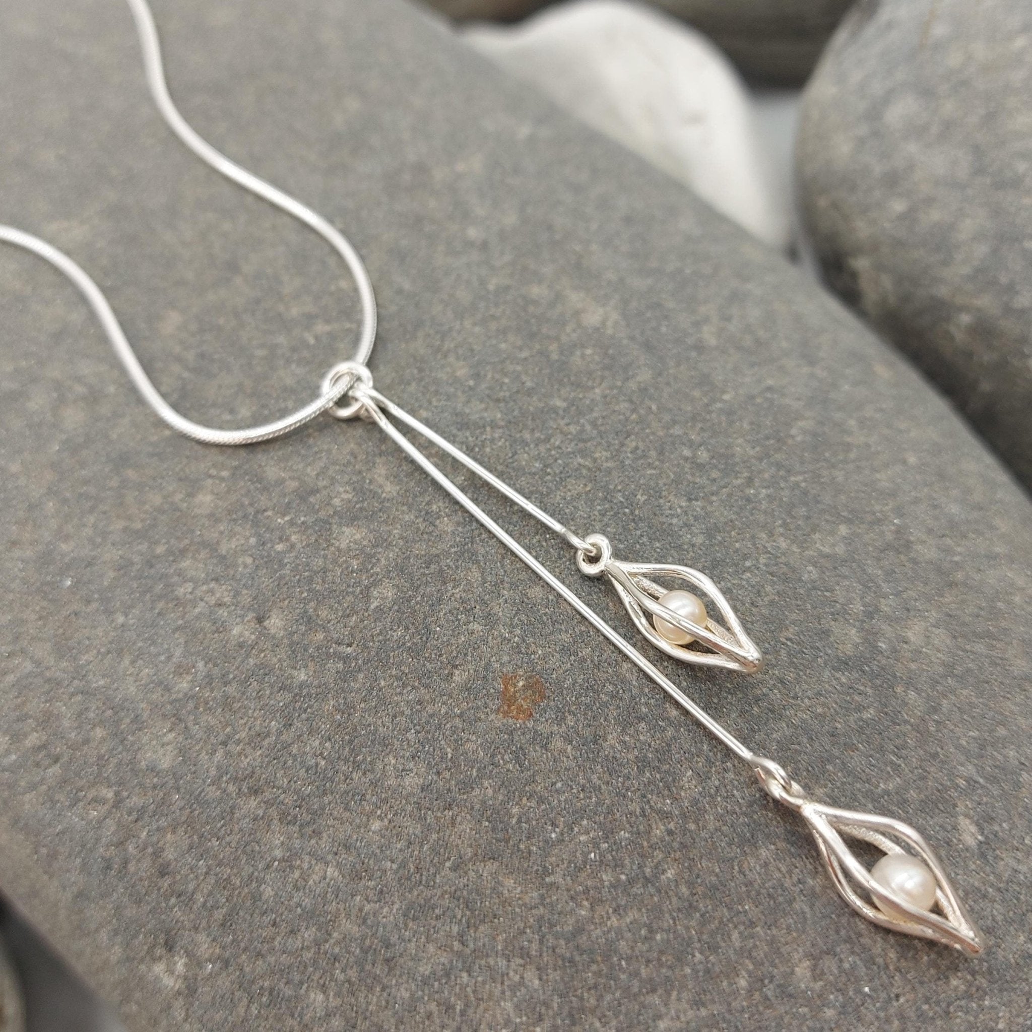 Sterling silver double drop white pearl necklace on a large grey rock. The necklace features two 925 sterling silver seed cage charms with one 5mm white freshwater cultured pearl inside each cage, each hanging from different length silver wire drops.