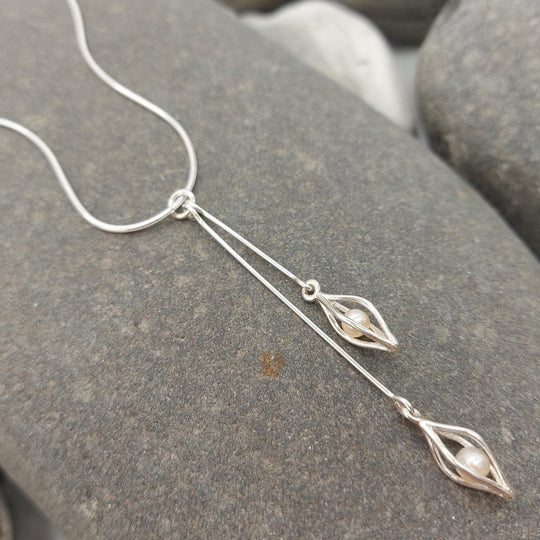 Sterling silver double drop white pearl necklace on a large grey rock. The necklace features two 925 sterling silver seed cage charms with one 5mm white freshwater cultured pearl inside each cage, each hanging from different length silver wire drops.