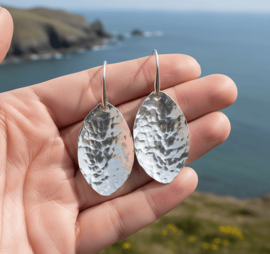 Close-up of pair of 5cm sterling silver hammered leaf earrings held in hand with Welsh coastal background