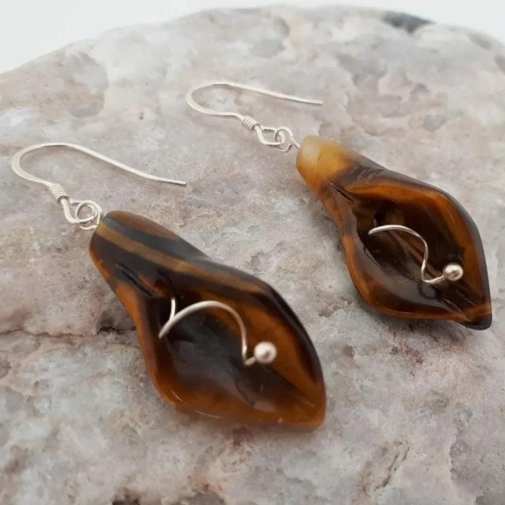 A pair of 925 sterling silver tigers eye calla lily earrings with shepherd hook closures, displayed on a rock.